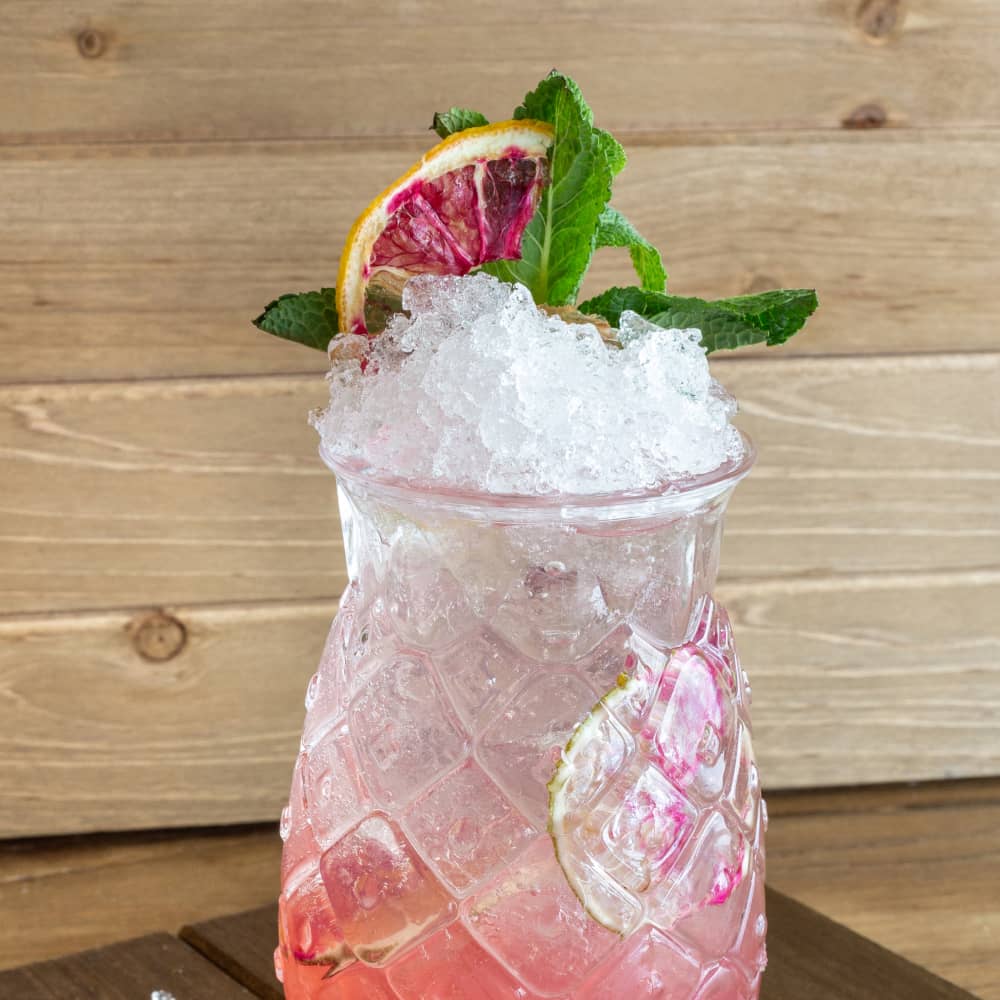Pink cocktail garnished with fresh fruit in a pineapple-shaped glass